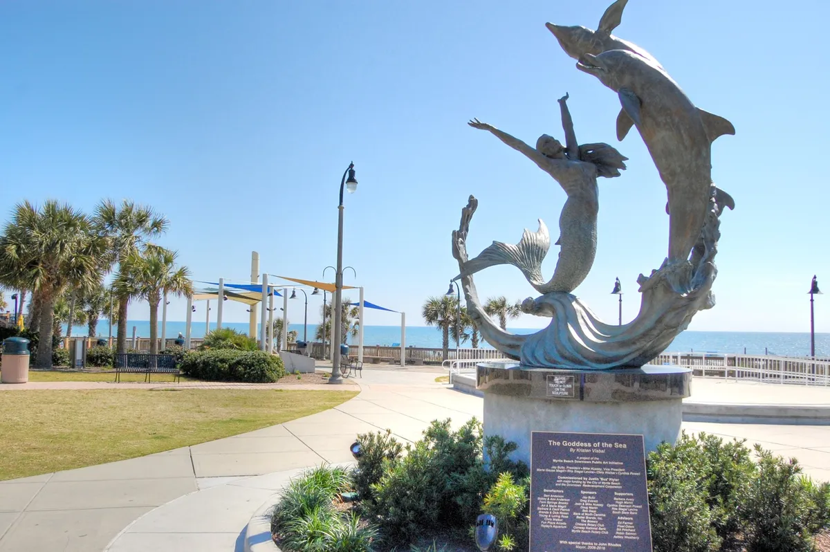 An inviting boardwalk lined with palm trees overlooking the ocean, featuring the striking statue of the Goddess of the Sea. The area is lively yet maintains a relaxed seaside vibe, perfect for romantic strolls or capturing special moments against the backdrop of the ocean.