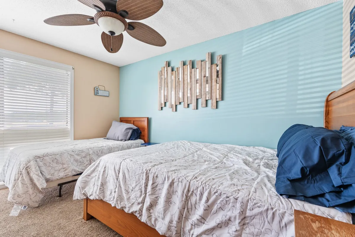 This interior image features two beds with patterned bedding, decorative wall art, a ceiling fan, and soft natural light filtering through blinds. The arrangement suggests space for multiple guests and a calming environment ideal for rest.