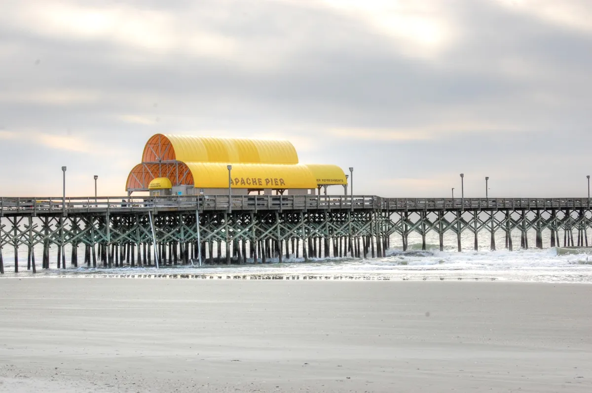 This image highlights the iconic Apache Pier, with its distinctive yellow-roofed wooden structure stretching out over the ocean. The scene features visitors strolling, fishing, and taking in the scenic seaside atmosphere, all under soft coastal light.