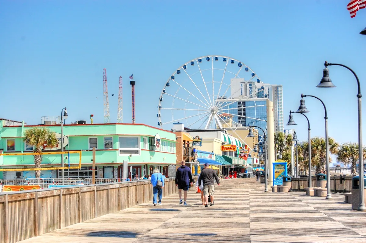 A vibrant boardwalk scene at sunset with a ferris wheel, colorful buildings, and groups of people walking, shopping, and enjoying the atmosphere.