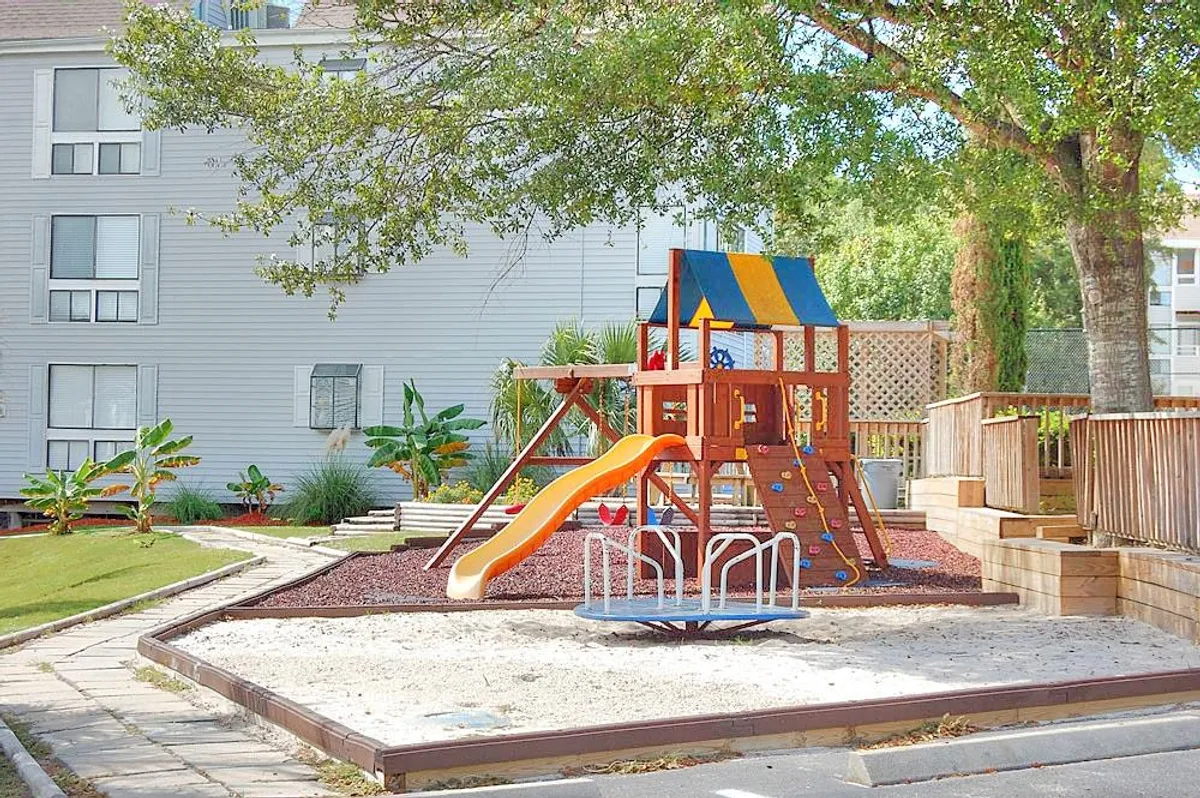 A cheerful playground surrounded by landscaped greenery, featuring slides and a merry-go-round. Children are actively playing and laughing, with families gathered for outdoor fun and picnics under sunny skies.