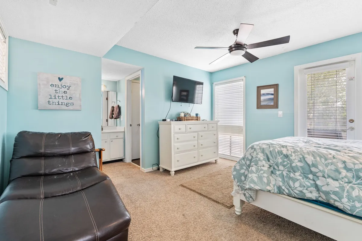 A cozy bedroom with a bed dressed in floral bedding, a comfortable leather chair, a white dresser, and a TV. The room is filled with morning light, inviting relaxation and quiet moments.