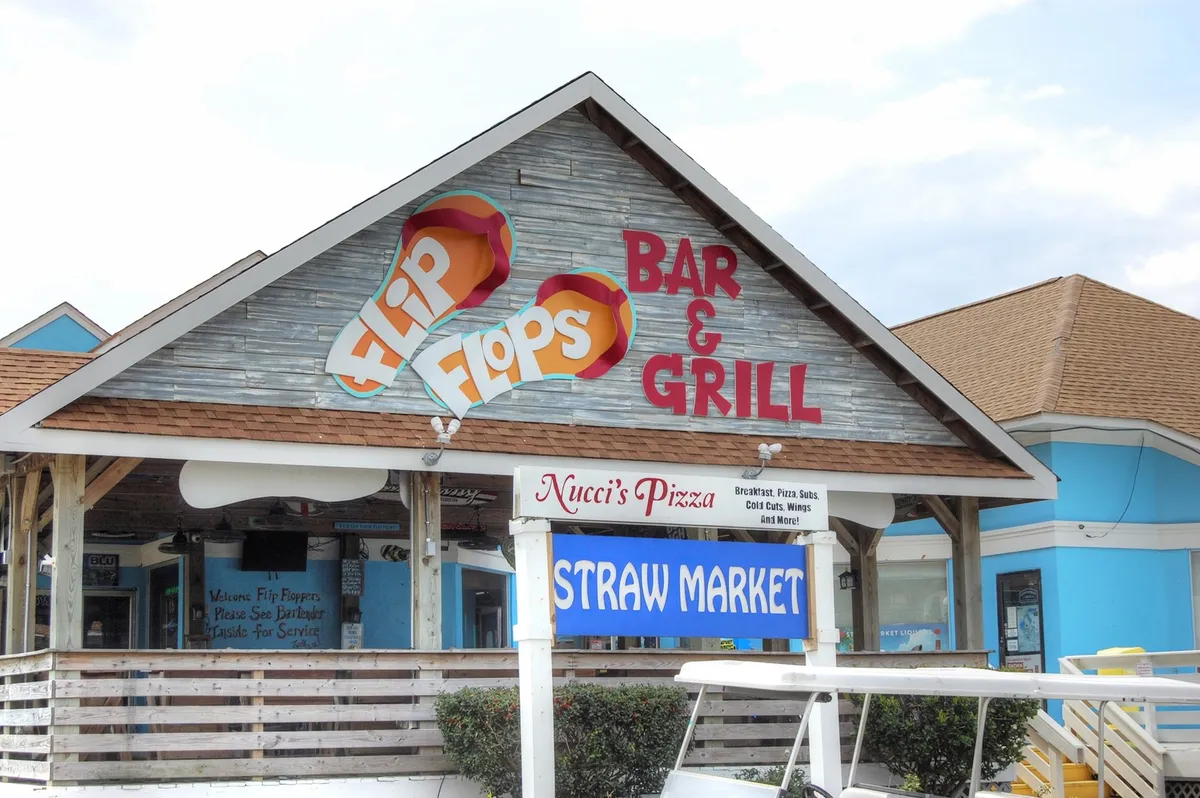 The exterior signage of Flip Flops Bar & Grill and Nucci's Pizza stands out, inviting guests to enjoy dining, socializing, and live music in a cheerful, beach-themed setting. The scene radiates casual fun and an easygoing, friendly atmosphere.