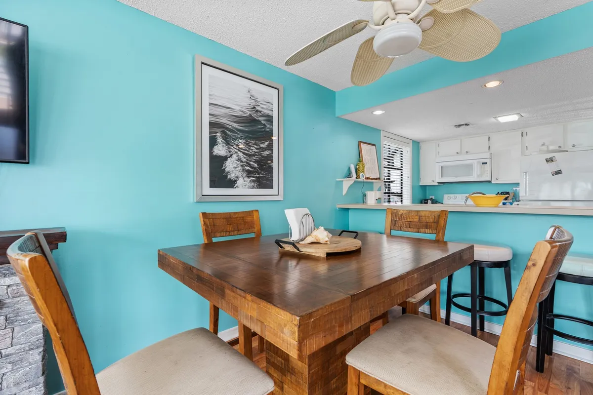 This image features a wooden dining table set against a turquoise wall, with a framed ocean photograph and a ceiling fan. The overall mood is welcoming and cheerful, emphasizing the communal aspect of dining and conversation.