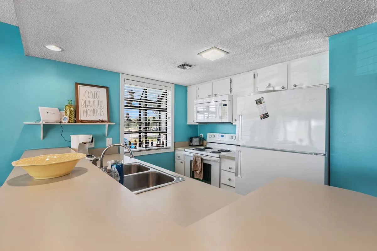 A bright kitchen space with a blue wall, kitchen island, window with blinds, and a cheerful decorative sign. The room is designed for cooking, entertaining, and gathering with family and friends.