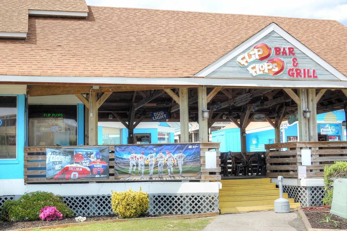 The exterior of Flip Flops Bar & Grill with colorful banners, outdoor seating, and groups of people socializing in a friendly, casual setting.