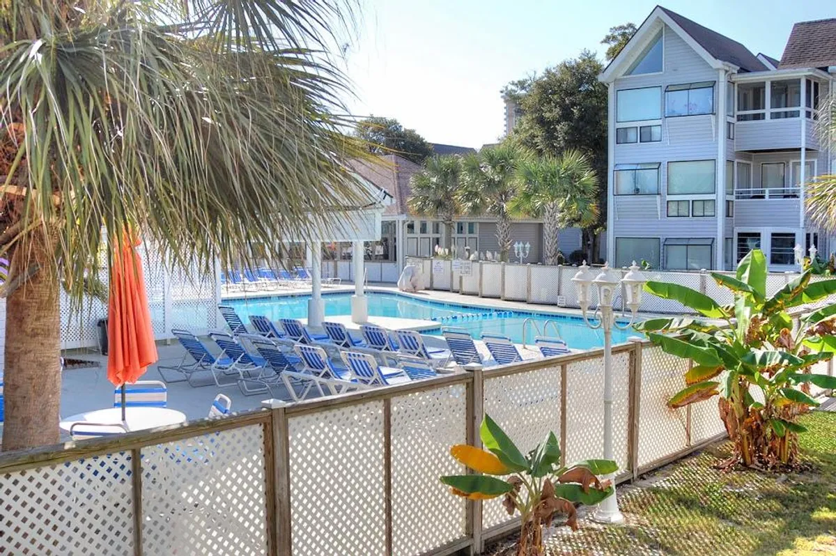 This image showcases a sparkling swimming pool surrounded by lounge chairs and palm trees, with an orange umbrella adding a pop of color. The building facade is visible in the background, creating a lively and inviting atmosphere suggestive of resort-style living.