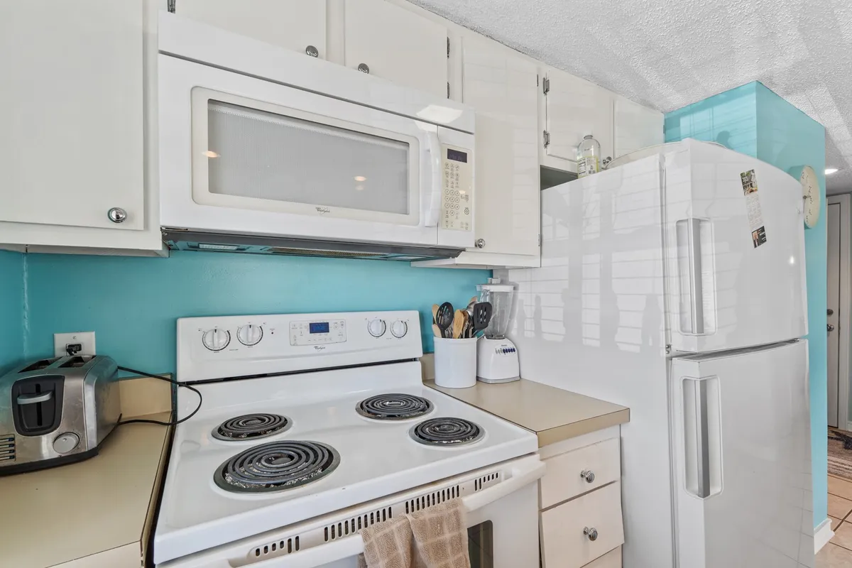 The image centers on a stove, microwave, and refrigerator against a turquoise backsplash, with a clean kitchen counter in view. The space looks organized and ready for cooking or gathering, supporting the idea of meal prep and group activities.