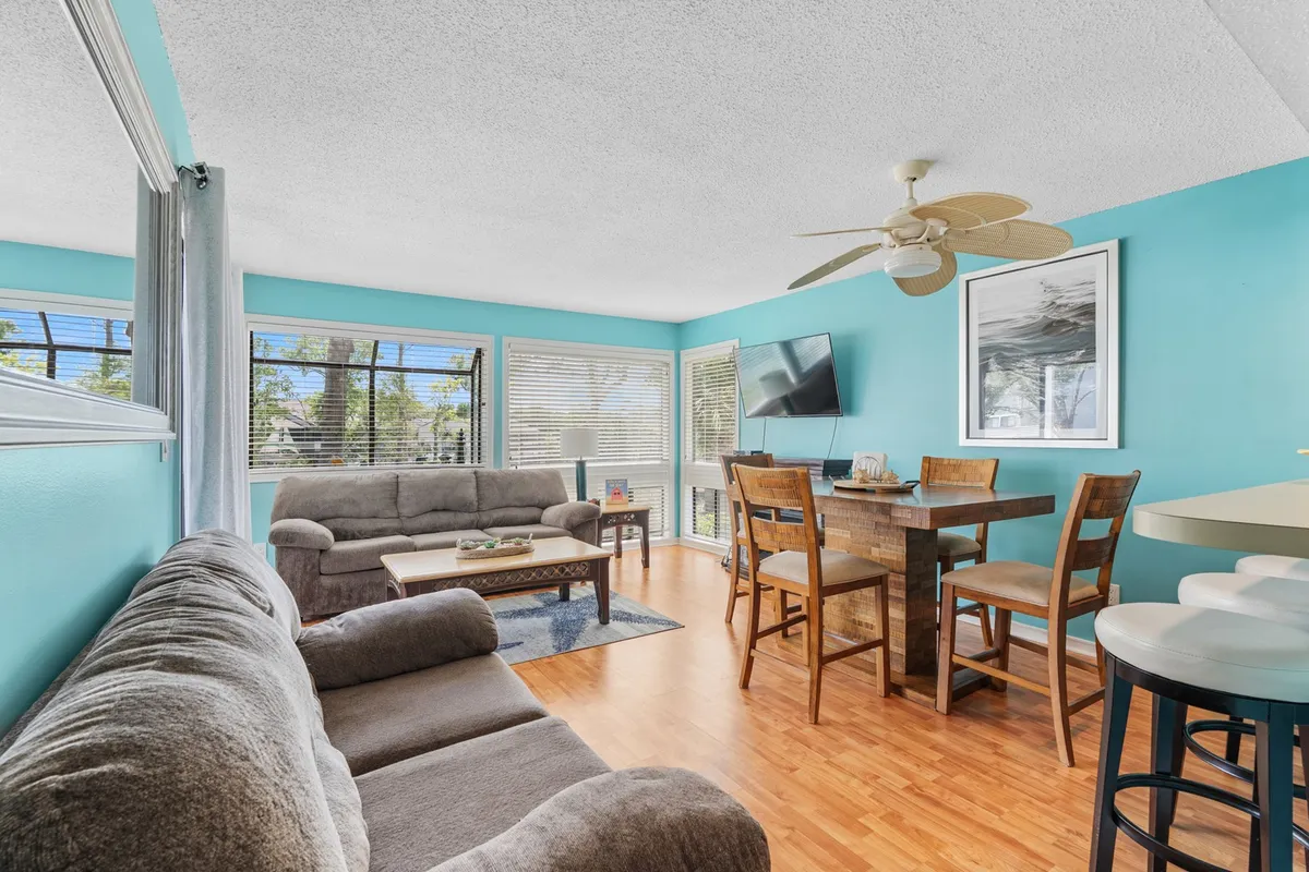 A spacious, sunlit living room featuring plush, comfortable sofas arranged for conversation, a large window flooding the space with natural light, a TV for entertainment, a ceiling fan for comfort, and an adjacent dining table—showcasing the heart of the home where guests can relax or connect.