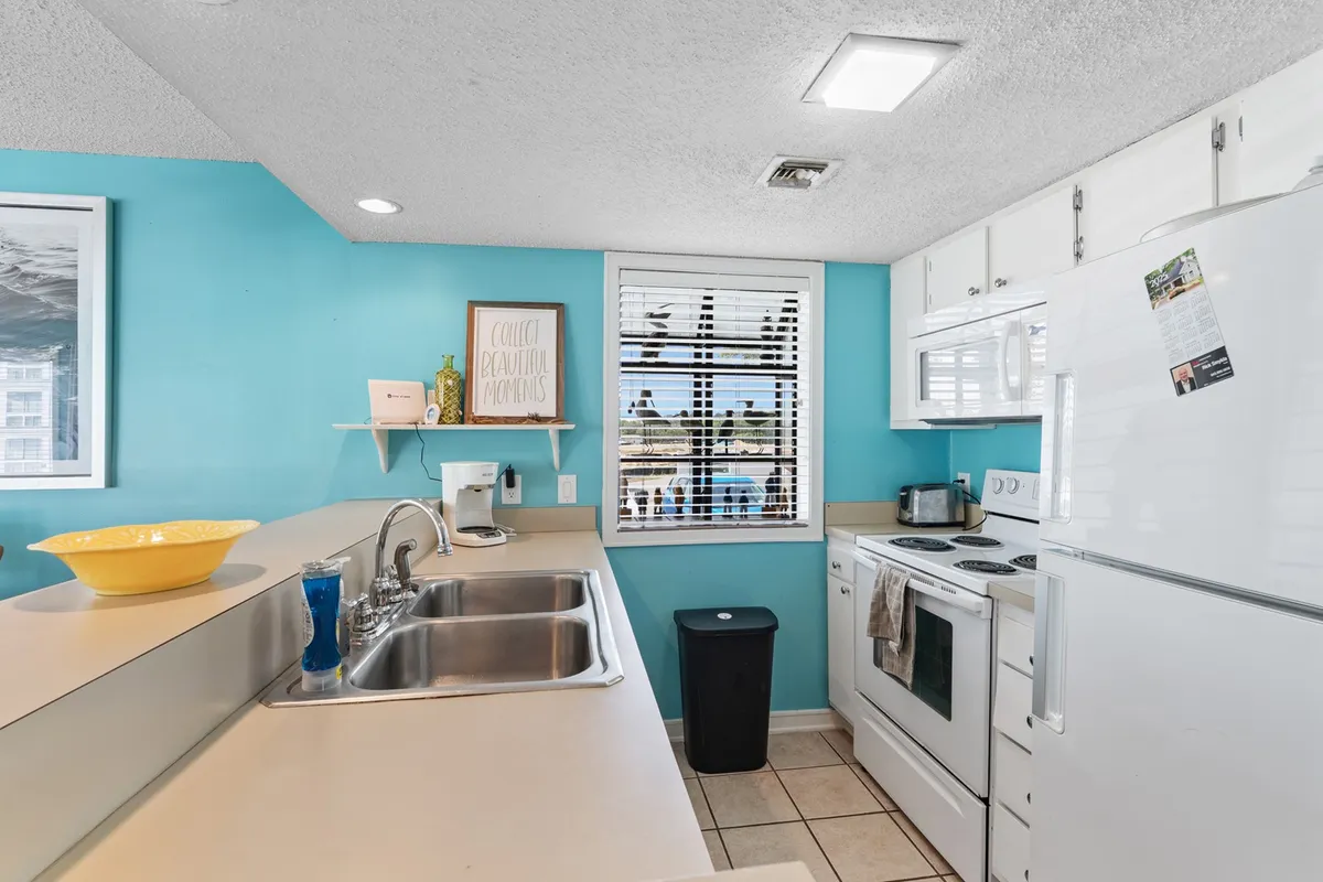 This image showcases a bright and cheerful kitchen area, with bright blue walls, modern appliances, a sink area, and a window with blinds. The scene is well-lit, highlighting the kitchen's full-size appliances and a welcoming atmosphere for meal preparation.